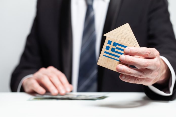 Businessman,Hand,Holding,Wooden,Home,Model,With,Greek,Flag.,Insurance Businessman hand holding wooden home model with Greek flag. insurance and property concept, source: Shutterstock
