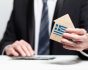 Businessman hand holding wooden home model with Greek flag. insurance and property concept, source: Shutterstock